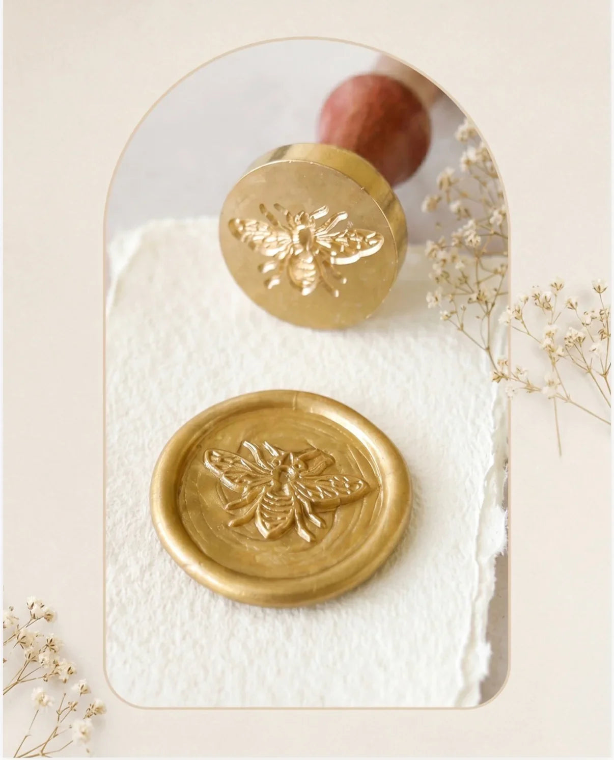 Close-up shot of a metallic gold wax seal stamped with an intricate honey bee impression, set on deckle-edged handmade paper. Above sits the brass seal head engraved with the same bee design, attached to a smooth wooden handle. The scene is framed with a soft arch, accented by delicate dried white flowers, from The Natural Paper Company.