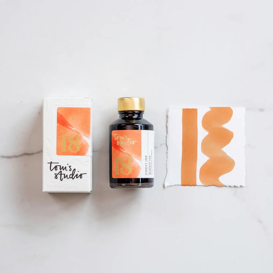 A bottle of orange fountain pen ink labeled 'Marmalade 18' by Toms Studio, accompanied by a sample of the ink used for calligraphy on paper with a gold-foiled label.