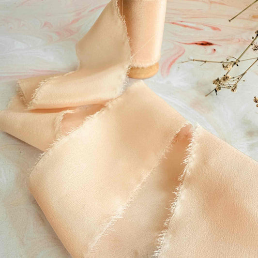 A peach colored silk ribbon with a raw edge, displayed next to a wooden spool and some dried flowers.