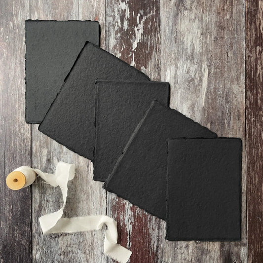 A collection of black handmade paper sheets with a textured surface and a deckled edge, displayed on a wooden background with a roll of white paper and a yellow object to the side.