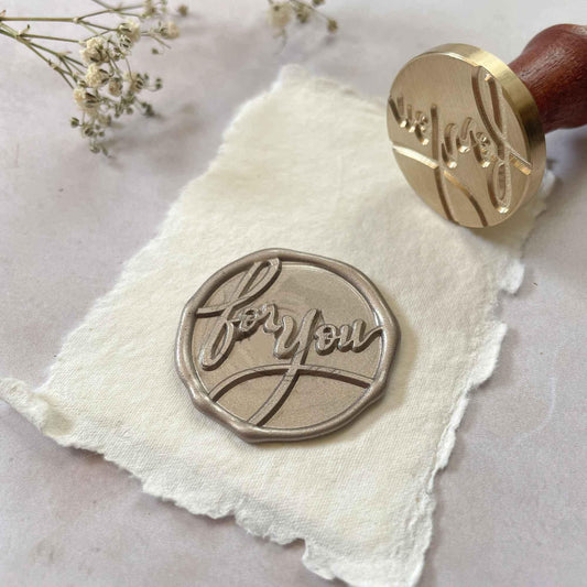 A brass wax stamp with a wooden handle, pressed into a wax seal on a piece of paper, displaying the inscription 'FOR YOU'.
