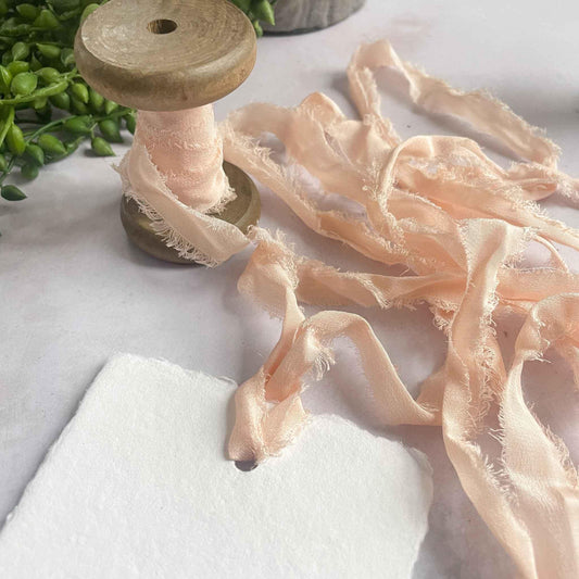 Rolls of silk ribbon in various shades of peach and pink, with a wooden spool and a needle, on a white surface.