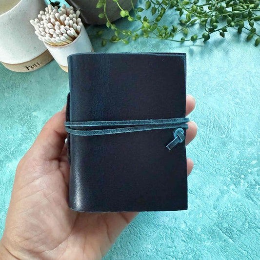 Hand holding a dark blue leather notebook with a turquoise strap against a light blue background.