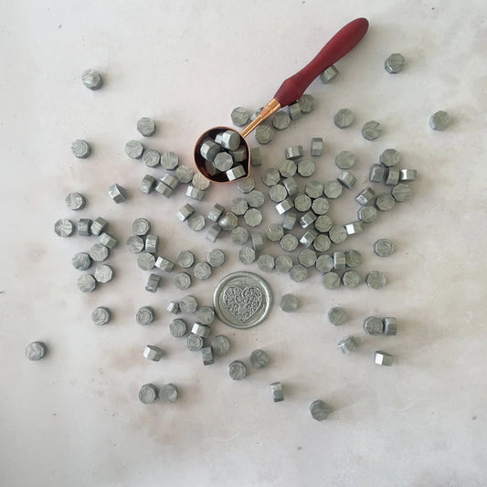 A collection of silver metallic sealing wax beads scattered on a surface with a few beads still attached to a red melting spoon.