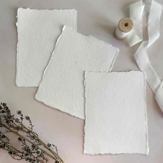 Stack of white handmade cotton rag envelopes with a deckled edge, placed on a similar white card, with a label tied to the stack displaying the brand name.
