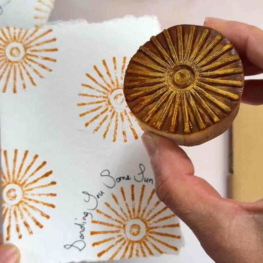 Hand holding a wooden stamp with sun design on a white paper with similar sun patterns.