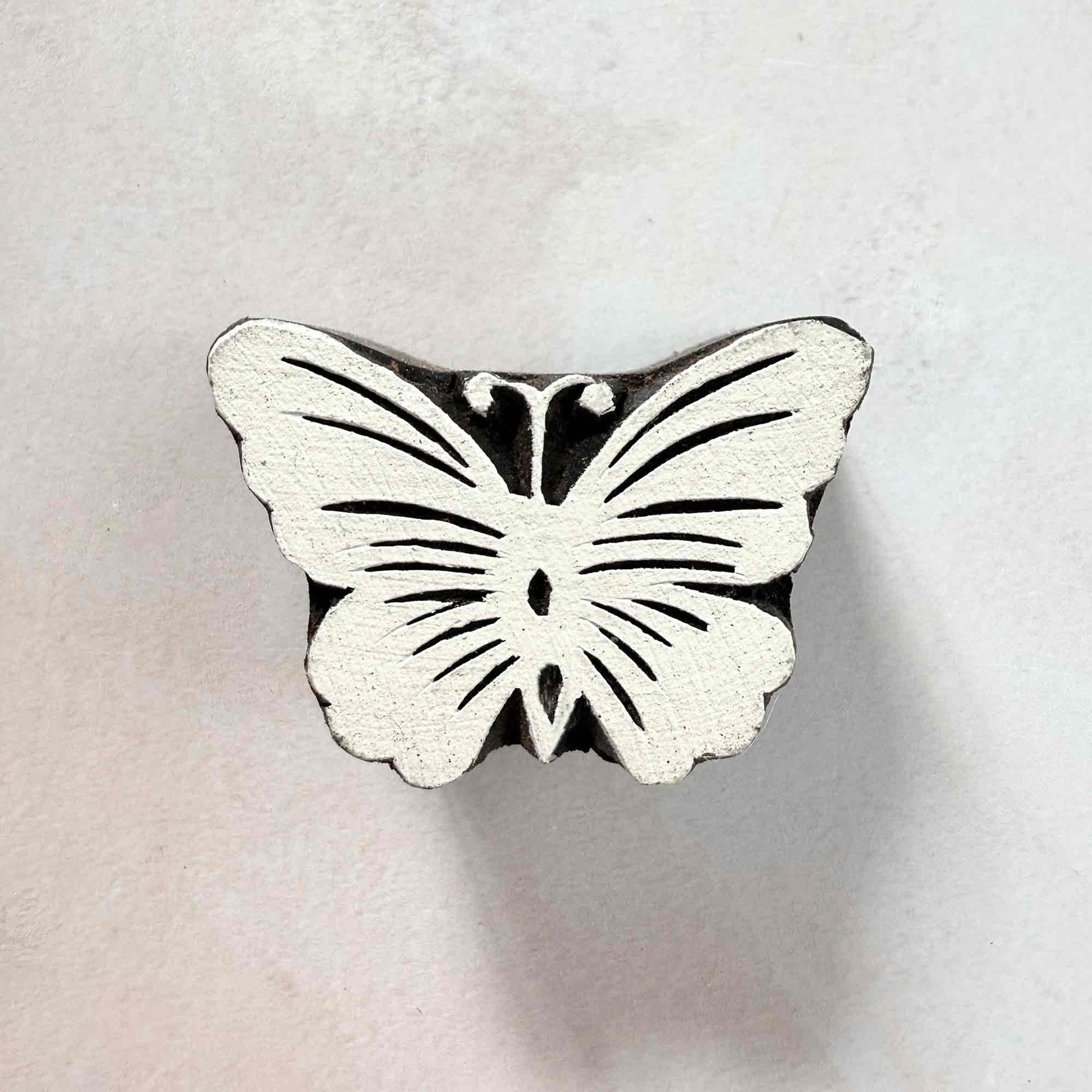 Indian wood block stamp — Flutter butterfly face-on view on soft pale background, symmetrical wings with fine curved veins, hand-carved wooden stamp, classic traditional printing block nature motif