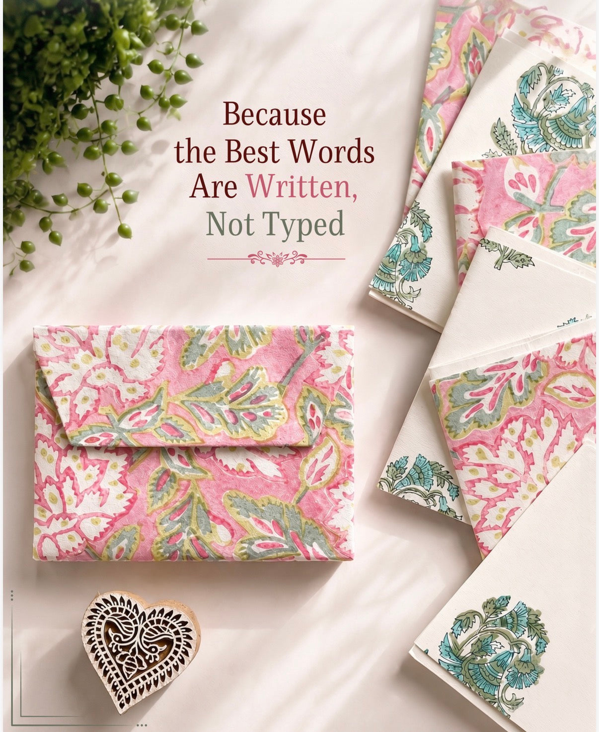 Flat-lay view of a handcrafted notecard set, hand block-printed with detailed floral motifs in pink, teal and green, crafted from recycled cotton paper. Displayed are printed envelopes, folded notecards, and an ornate wooden heart printing block. Overlaid text: “Because the Best Words Are Written, Not Typed”. Warm natural styling with greenery, from The Natural Paper Company.