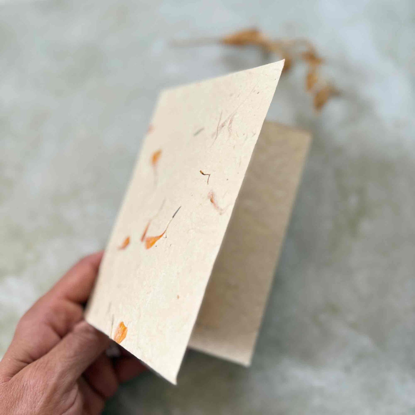 Hand holding an open folded lokta paper card, highlighting its thick textured surface and embedded dried marigold petals. Eco-friendly, tree-free handmade stationery.