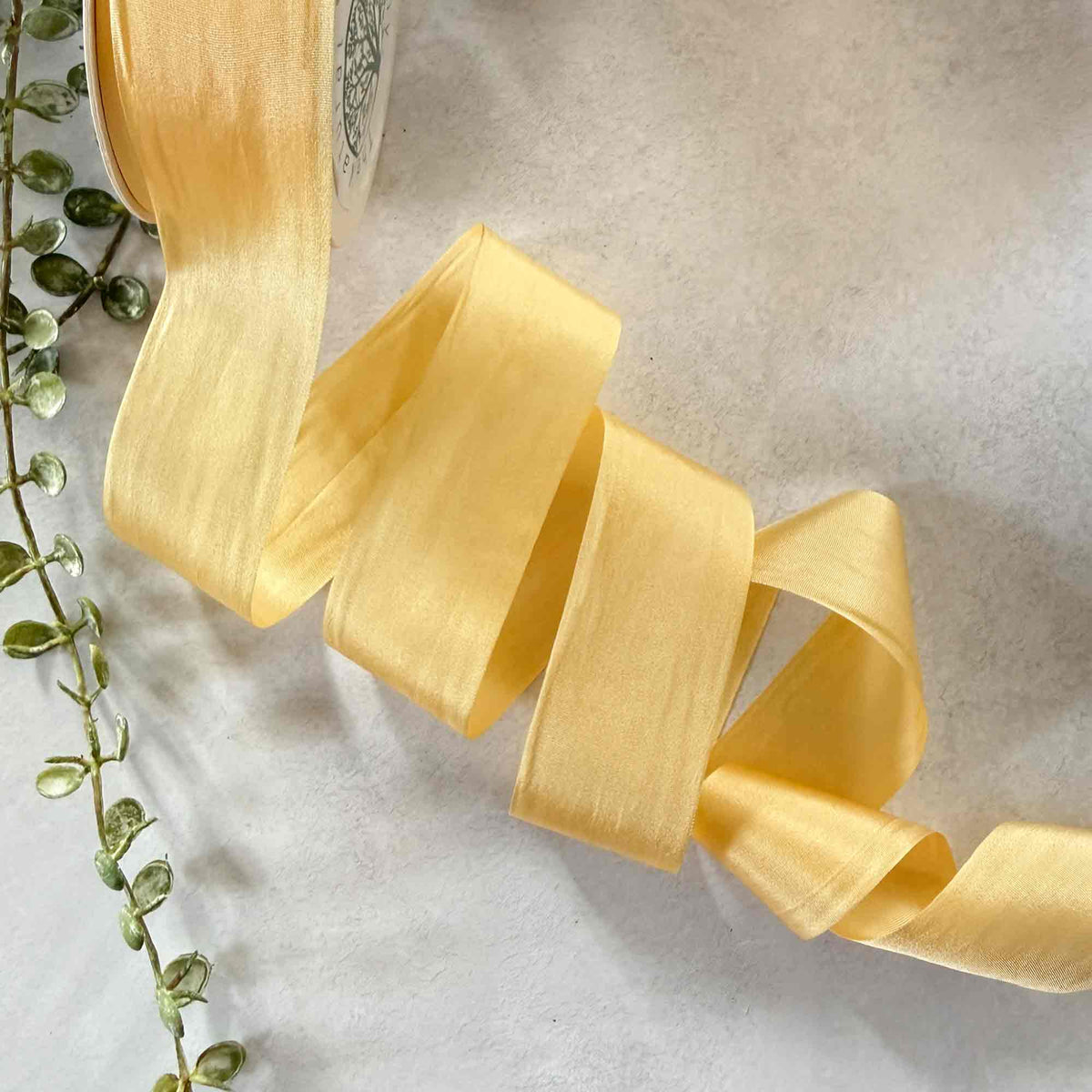 Wide Buttermilk Habotai Silk Ribbon styled with greenery, showing its soft creamy yellow colour and silky finish.