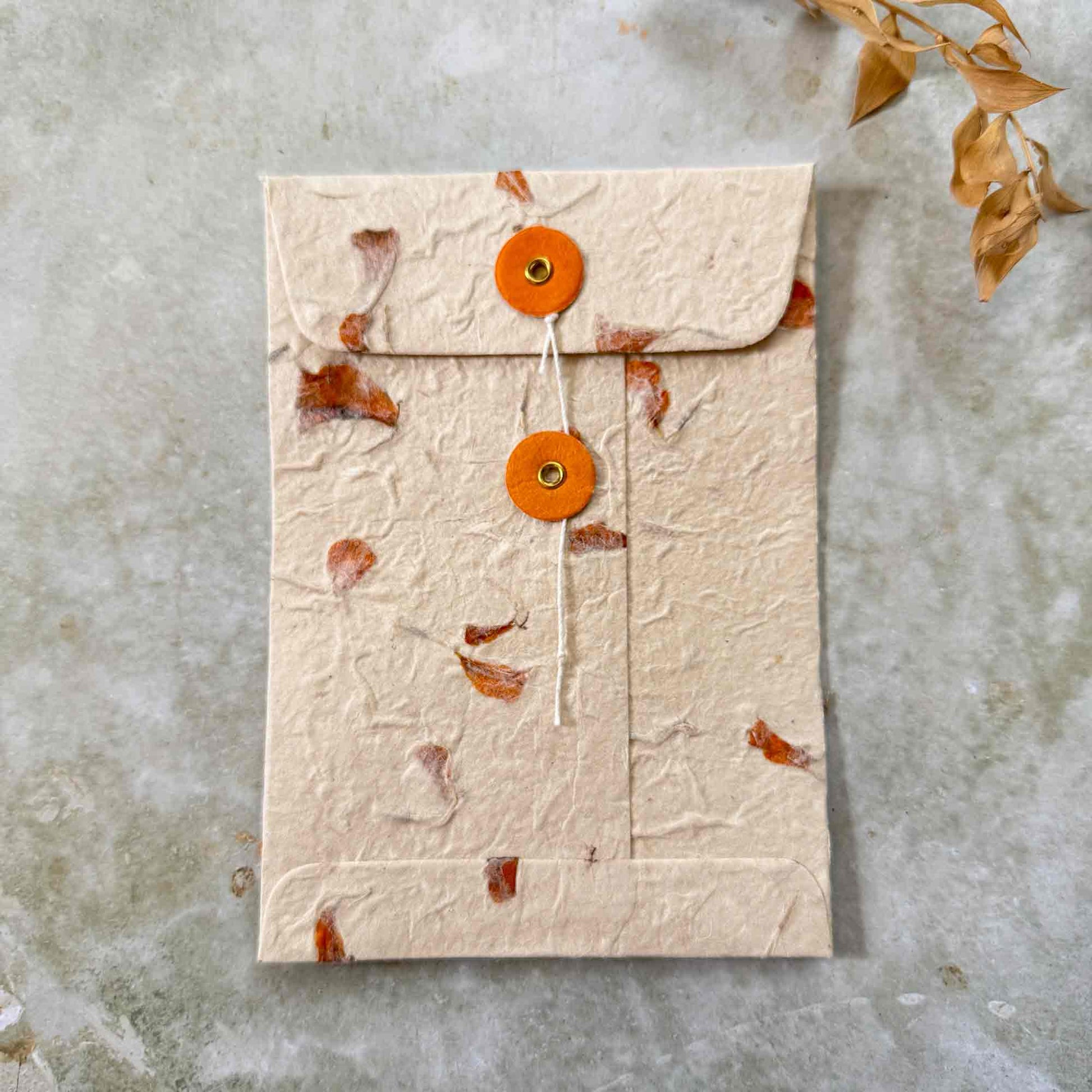 Closed handmade lokta paper envelope with matching orange buttons and cotton string tie, featuring embedded dried orange marigold petals. Sustainable zero-tree stationery.