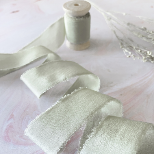 silk ribbon in light sage green colour.  Luxury habotai silk ribbon on a wooden spool.  Dusky sage green silk ribbon with a frayed edge.  Perfect ribbon for decorating wedding stationery
